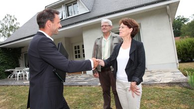 Drei Personen stehen vor einem Haus, ein Mann im Anzug reicht einer Frau die Hand. 