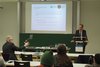 Der Raum von hinten. Volle Reihen. Vorne steht Prof. Dr. Kube am Rednerpult vor seiner PowerPoint. Prof Dr. Raab stellt aus der ersten Reihe eine Frage. 