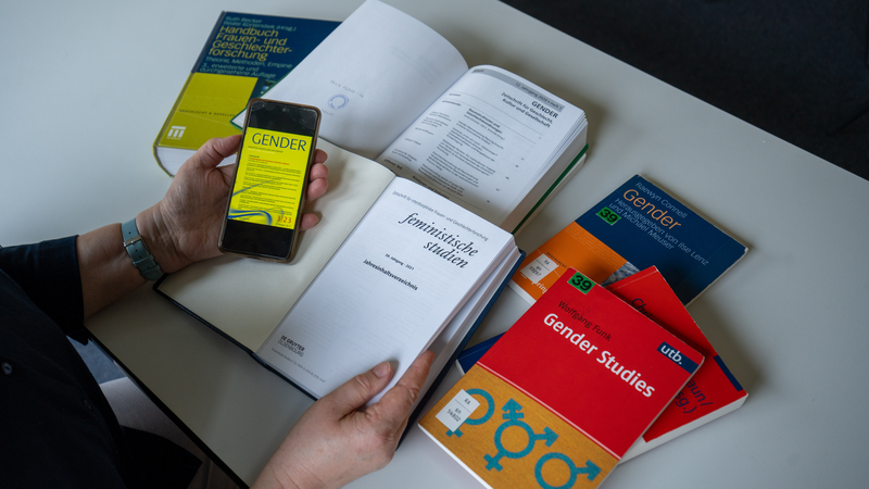 Symbolbild: Frauenhand, hält Smartphone auf dem Bildschirm zu lesen Gender, im HIntergrund aufgeschlagene Literatur zum Thema Geschlechterstudien