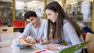 Two persons sitting at a table, looking in a book and taking notes.