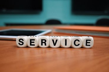 Auf einem Tisch stehen sieben Würfel nebeneinander, auf diesen sind jeweils ein Buchstabe. Gemeinsam ergeben dieses das Wort Service. Im Hintergrund ist ein Tablet und ein Buch.