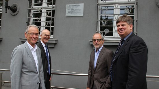 Dr. Jürgen Brauer, Peter Fritzen, Klaus Jensen und Dr. Thomas Grotum vor dem grauen Gebäude der Christophstraße 1 die Gedenktafel aus Glas hängt an der Außenwand