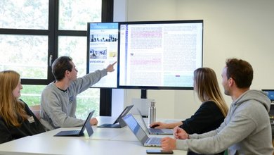 Vier Studierende diskutieren vor einem großen Monitor