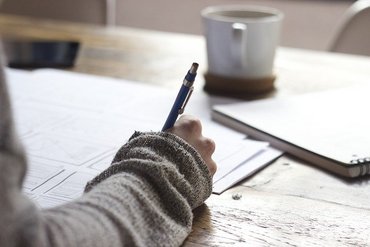 Eine Hand einer Person mit einem Kugelschreiber, die auf mehreren weißen Blättern schreibt. Im Hintergrund ist ein Notizbuch sowie eine Tasse.