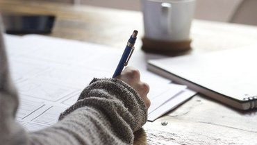 Eine Hand einer Person mit einem Kugelschreiber, die auf mehreren weißen Blättern schreibt. Im Hintergrund ist ein Notizbuch sowie eine Tasse.