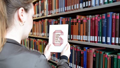 Frau hält das Grundgesetz in der Hand vor einem Bücherregal in der Bibliothek