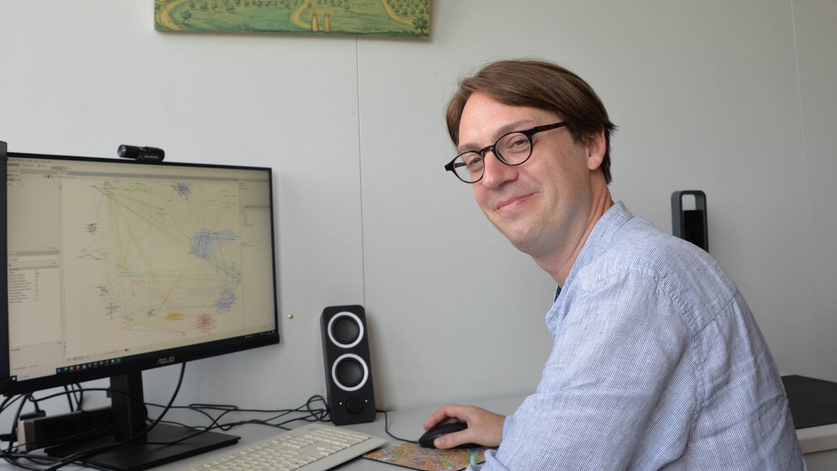 Prof. Dr. Damien Tricoire sitzt am Rechner und schaut in die Kamera