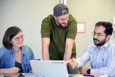 Drei Personen sitzen vor Laptop, eine redet und zeigt auf den Bildschirm.