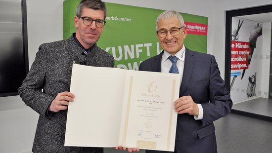 Prof. Jäckel hält die Urkunde über die Ehrennadel der Handwerkskammer in der Hand, daneben Rudi Müller, Präsident der Handwerkskammer Trier. 