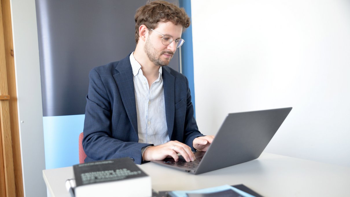 [Translate to Englisch:] Marc Seul vor Laptop, Bücher auf dem Tisch