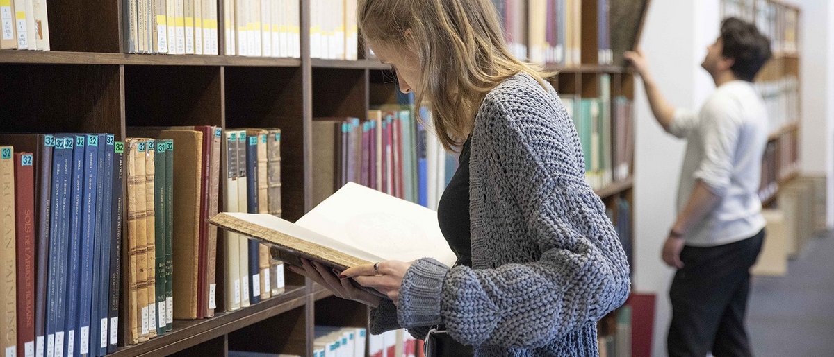 Studierende vor einem Bücherregal stehend und in der Hand ein Buch lesend