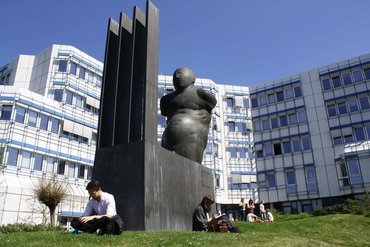 Studierende sitzen auf Wiese an einer Statue vor Gebäude D.