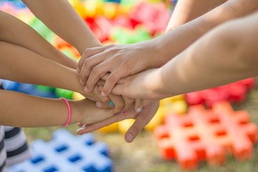 Sieben Kinderhände werden übereinandergehalten vor einem bunten Hintergrund.