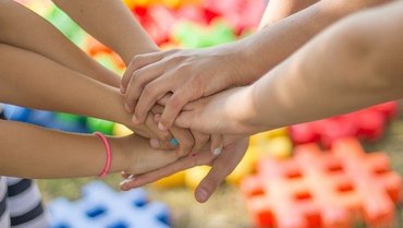 Sieben Kinderhände werden übereinandergehalten vor einem bunten Hintergrund.