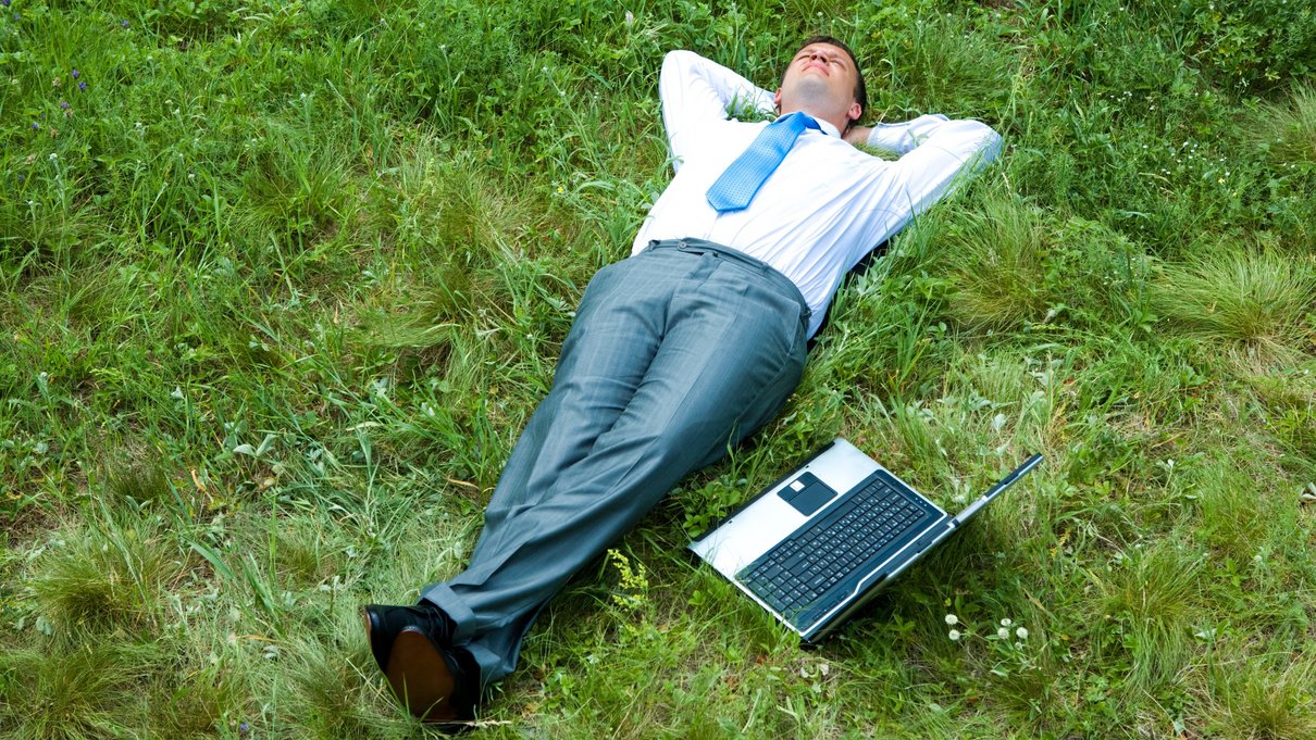 Mann liegt auf der Wiese neben einem Laptop und entspannt.