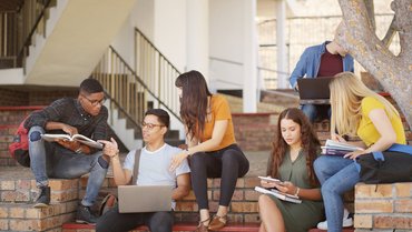 junge Erwachsene sitzen auf einer Mauer und einer zugehörigen Bank und tauschen sich vor Büchern, Laptops und Tablets miteinander aus