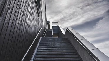 Eine Treppe, die gerade aus nach oben führt. Links ist die Wand eines Gebäudes und rechts ein bewölker Himmel.