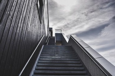 Eine Treppe, die gerade aus nach oben führt. Links ist die Wand eines Gebäudes und rechts ein bewölker Himmel.