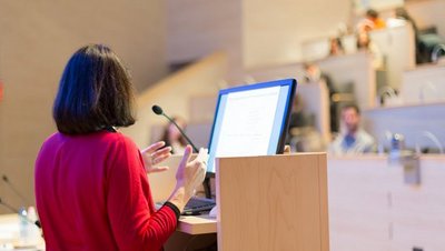 [Translate to Englisch:] Symbolfoto einer Professorin, sie steht am Rednerpult vor dem Hörsaalpublikum