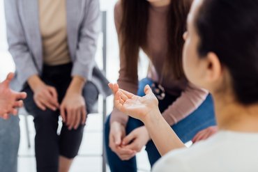 Blick über die Schulter einer Person, die mit der rechten Hand eine redebegleitende Geste macht, im Hintergrund zwei Personen die der ersten Person zugewand sitzen.