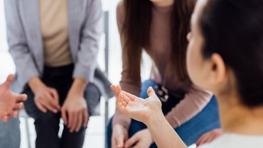 Blick über die Schulter einer Person, die mit der rechten Hand eine redebegleitende Geste macht, im Hintergrund zwei Personen die der ersten Person zugewand sitzen.