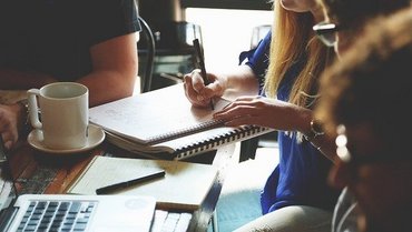 Vier Personen sitzen an einem Tisch. Eine Person schreibt in einen Block. Auf dem Tisch stehen eine weiße Kaffeetasse sowie ein Laptop.