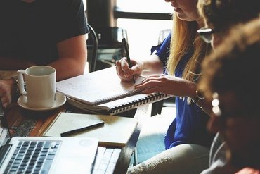 Vier Personen sitzen an einem Tisch. Eine Person schreibt in einen Block. Auf dem Tisch stehen eine weiße Kaffeetasse sowie ein Laptop.