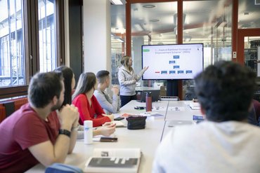 Studierende sitzen am Tisch und schauen zu Studentin, die einen Vortrag hält.