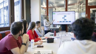 Studierende sitzen am Tisch und schauen zu Studentin, die einen Vortrag hält.