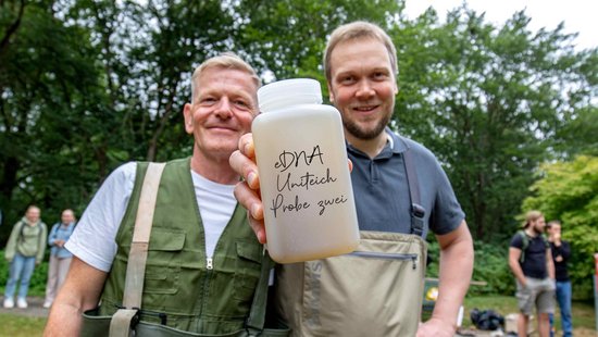 [Translate to Englisch:] Zwei Forscher der Uni Trier halten eine Gewässerprobe aus dem Uni Teich in die Kamera 