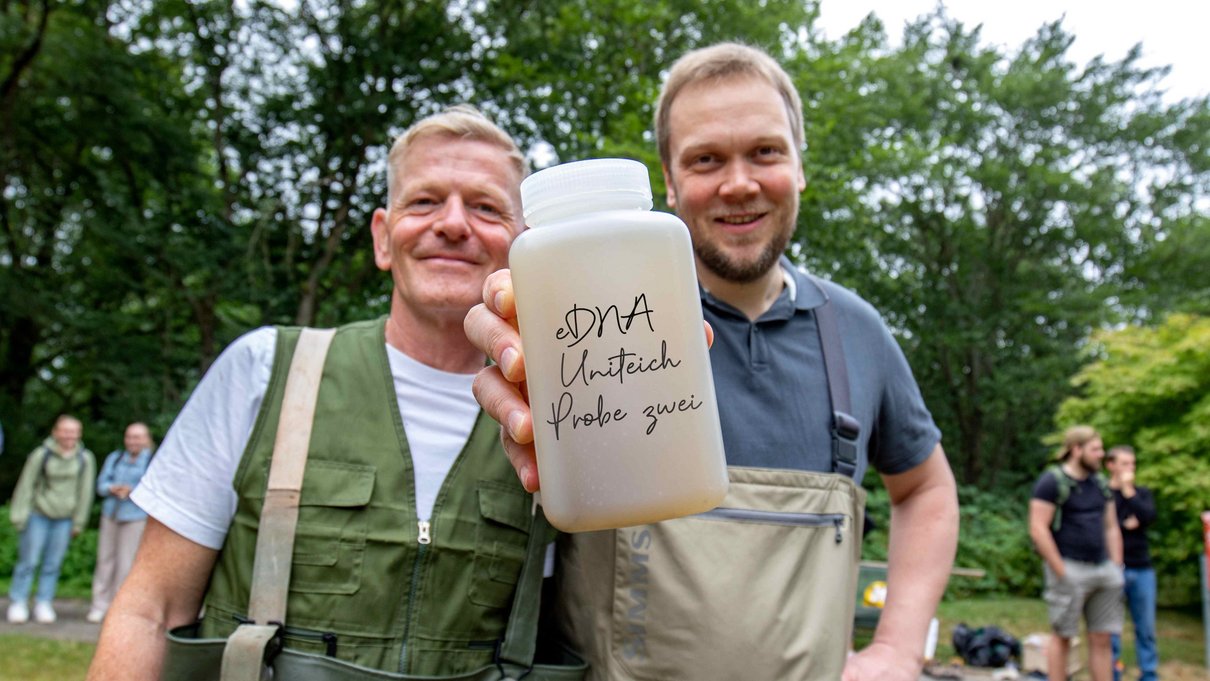 Zwei Forscher der Uni Trier halten eine Gewässerprobe aus dem Uni Teich in die Kamera 