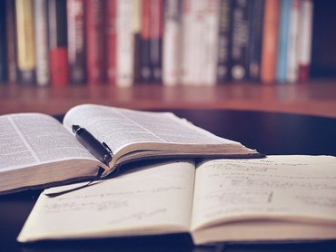 Zwei aufgeschlagene Bücher liegen auf einem Tisch vor einem Bücherregal mit weiteren Büchern.
