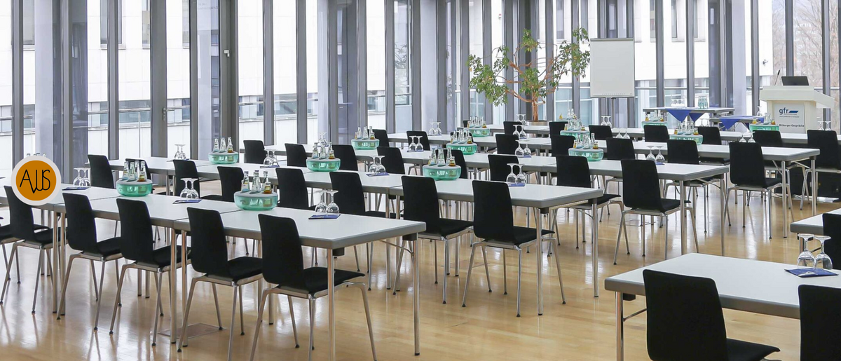 The picture shows a large, modern conference room with several rows of tables and chairs arranged in a seminar style. There are bottles of water and glasses on the tables, and in the background you can see a flipchart and a window with a view outside.