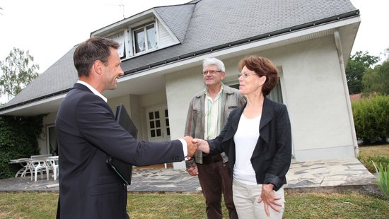 Drei Personen vor einem Haus. Mann mit Anzug reicht einer Frau die Hand