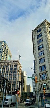 This picture shows part of the Exchange District in Manitoba. You can see some higher buildings with a lot of windows and part of a busy street and red lights. The sky is blue with some clouds but it is sunny.