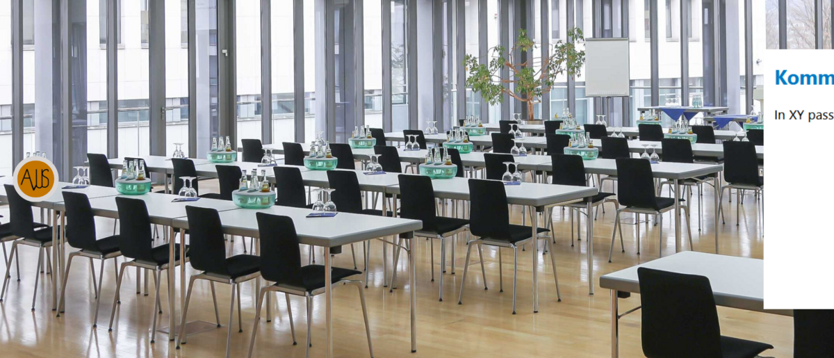 The picture shows a large, modern conference room with several rows of tables and chairs set up in a seminar style. There are water bottles and glasses on the tables, and in the background you can see a flipchart and a window with a view outside. 