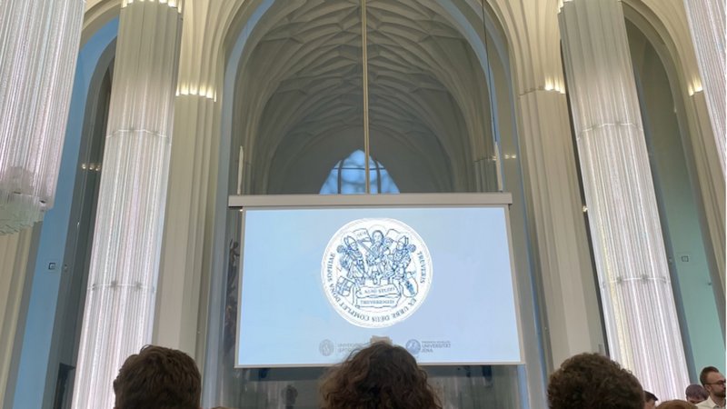 The picture shows the interior of a modern hall with high, bright columns and a projection on a screen. The University of Trier's coat of arms can be seen on the screen. People in the audience can be seen in the lower section. At the bottom left is a blue box with the inscription “EVENTS & NEWS”