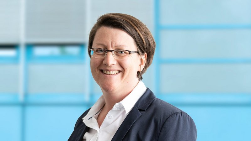 The picture shows the smiling chairwoman in a professional outfit against a blurred background in shades of blue. In the bottom left corner is a blue box with the inscription “PROFESORSHIP"”