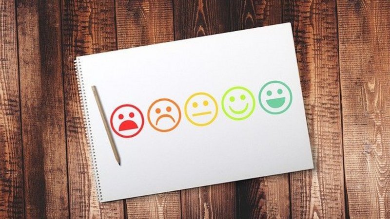 [Translate to Englisch:] Ein Blatt Papier mit fünf gemalten Smileys in verschiedenen Farben, von rot und unglücklich auf der linken Seite bis zu dunkelgrün und sehr glücklich auf der rechten Seite des Papiers. Als Hintergrund sieht man Holzdielen.