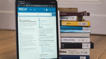 Tablet zeigt Bibliotheksseite, im Hintergrund sind mehrere Bücher gestapelt.