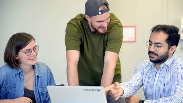 Drei Personen sitzen vor Laptop, eine redet und zeigt auf den Bildschirm.